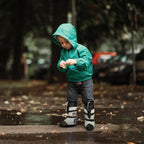 Toddler in green rain jacket wearing astronaut boots on wet outdoor pavement